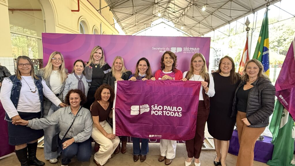 A imagem mostra um grupo de mulheres em pé, sorrindo para a câmera. Elas estão segurando uma faixa roxa com o logotipo e o slogan "São Paulo por Todas". As mulheres parecem ser funcionários públicos ou representantes de uma organização, dado o contexto da imagem e a faixa que carregam. Há bandeiras ao fundo, incluindo a brasileira, sugerindo um evento oficial ou político. As mulheres estão vestidas de forma profissional, com roupas em tons neutros e alguns acessórios. O ambiente parece ser uma área coberta, possivelmente um corredor ou hall de um prédio público. A imagem transmite uma sensação de unidade e coletividade.