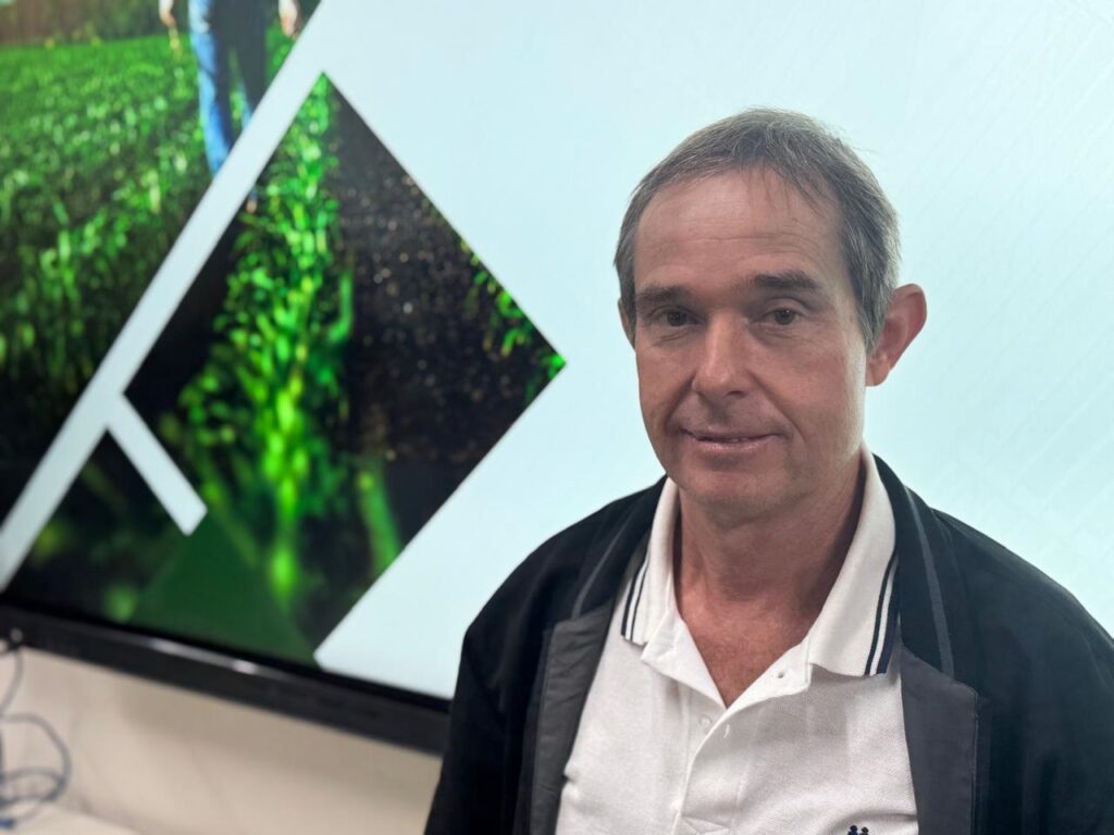 A imagem mostra um homem de meia-idade, com cabelos grisalhos curtos e pele clara. Ele está vestindo uma camisa polo branca com detalhes em azul marinho na gola e nas mangas, sobre a qual usa um casaco preto com detalhes em cinza. O homem está olhando diretamente para a câmera com uma expressão neutra e um leve sorriso. Ao fundo, há uma tela grande exibindo uma imagem desfocada de um campo verde com plantas crescendo, possivelmente uma plantação. Há também formas geométricas brancas sobrepostas à imagem de fundo, que parecem fazer parte de um design gráfico. A iluminação parece ser artificial, com a luz vindo da frente do homem, iluminando seu rosto.