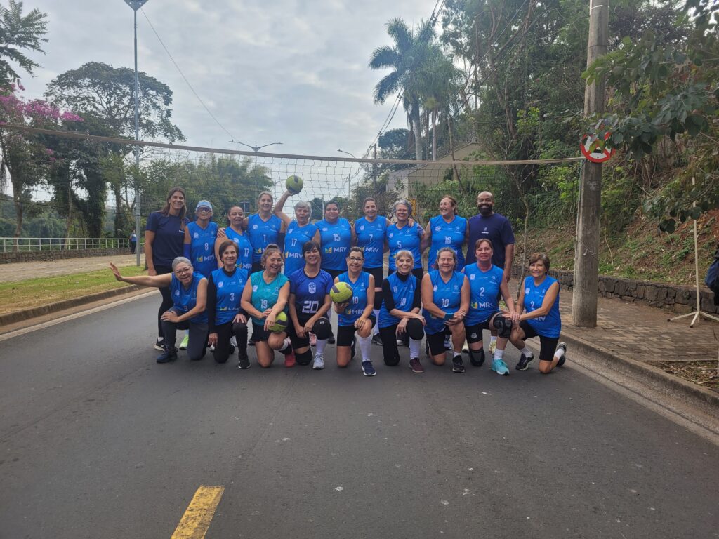 A imagem mostra um grupo de mulheres e um homem, aparentemente um time de vôlei, posado para uma foto em frente a uma rede de vôlei. As mulheres vestem uniformes azuis com números e o logo "MRV" visível em alguns uniformes. Elas estão sorrindo e algumas estão segurando bolas de vôlei. O homem está posicionado atrás das mulheres, vestindo uma camiseta escura. O fundo é uma rua e uma área arborizada. A cena sugere um evento esportivo ou um treino de vôlei ao ar livre. A imagem transmite um clima de camaradagem e alegria. O número de pessoas na imagem é de 17, incluindo o homem. A qualidade da imagem é boa, permitindo a identificação clara de rostos e detalhes dos uniformes. A iluminação parece ser natural, provavelmente em um dia ensolarado. A composição da foto é bem equilibrada, com o grupo centralizado e o fundo proporcionando contexto.