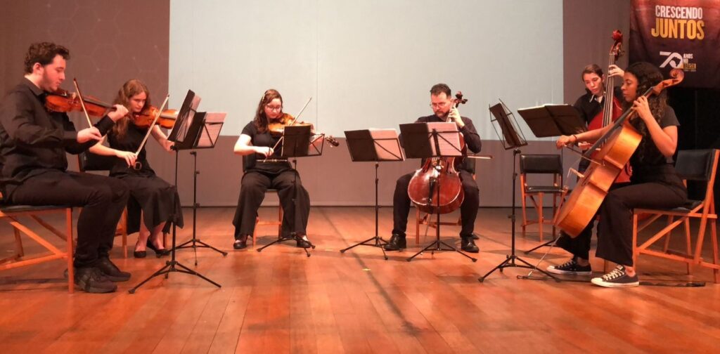 A imagem mostra um grupo de seis músicos tocando instrumentos de corda em um palco. Todos os músicos estão vestidos com roupas pretas. À esquerda, dois músicos tocam violino, sentados em bancos baixos. O músico da frente tem cabelo cacheado e está tocando com o arco em seu violino. A musicista atrás dele tem cabelo longo e ondulado e também está tocando violino. No centro, uma musicista toca violino, sentada em um banco mais alto. Ela usa óculos e está concentrada em sua partitura. Ao lado dela, um músico toca violoncelo, sentado em um banco. Ele também está usando óculos e tem uma barba. À direita, um músico mais jovem toca violoncelo, em pé. Ele está vestido com uma camisa preta e calças. Ao lado dele, uma musicista toca violoncelo, sentada. Ela tem cabelo cacheado e está usando tênis pretos. Atrás dos músicos, há um telão branco com um padrão geométrico em tons de cinza e um banner com a inscrição "CRESCENDO JUNTOS" e o número "70" em destaque, indicando uma celebração de 70 anos. O palco é de madeira clara e a iluminação cria um ambiente de apresentação.