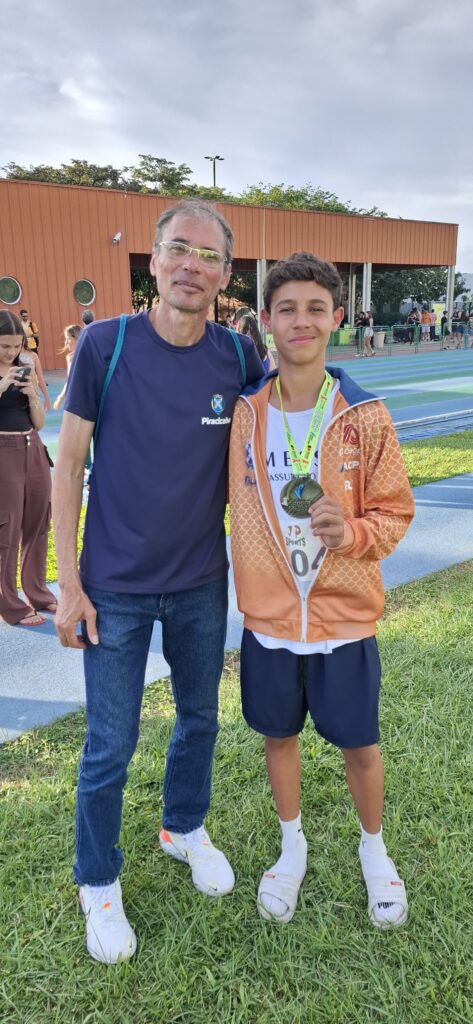 Na imagem, um homem mais velho e um jovem estão posando para uma foto em um dia ensolarado, provavelmente após um evento esportivo. O homem, que usa óculos e uma camiseta azul com o logo "Piracicaba", está com a mão no ombro do jovem. O jovem, por sua vez, veste uma jaqueta laranja sobre uma camiseta branca com o número "04" e exibe orgulhosamente uma medalha no pescoço, com a inscrição "Jogos Escolares". Ambos sorriem para a câmera, com o fundo mostrando uma pista de atletismo e uma estrutura de edifício com detalhes em laranja. A grama verde e o céu nublado completam a cena.