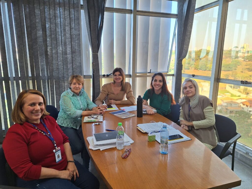 A imagem mostra cinco mulheres sentadas em torno de uma mesa de madeira numa sala de reuniões com grandes janelas que oferecem uma vista panorâmica de uma cidade. As mulheres parecem estar num encontro de trabalho ou numa reunião, com papéis, canetas e garrafas de água na mesa. Elas estão vestidas de forma profissional e parecem estar envolvidas numa conversa ou discussão. A sala é bem iluminada e tem um ambiente limpo e organizado. A atmosfera parece ser calma e profissional. As mulheres parecem ser colegas de trabalho ou membros de uma equipa, e a imagem sugere uma reunião produtiva e colaborativa.