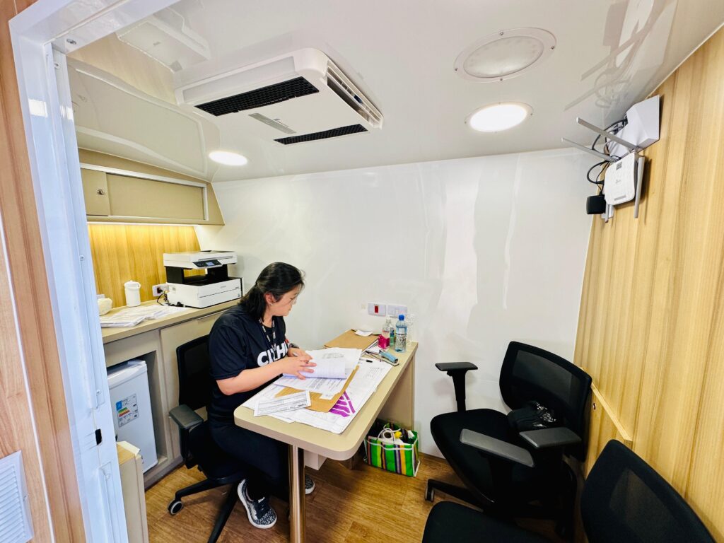 A imagem mostra um escritório compacto e funcional, onde uma pessoa trabalha concentrada em uma mesa repleta de papéis, cadernos e itens de papelaria. Ao fundo, há uma bancada com impressora, caneca e outros objetos organizados, sugerindo um ambiente de trabalho ativo. O espaço tem piso e paredes de madeira, teto branco com luminárias circulares e um aparelho de ar-condicionado. Duas cadeiras pretas completam o mobiliário, e há um roteador com cabos visíveis na parede lateral.