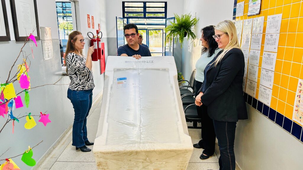 A imagem mostra um grupo de pessoas em um corredor de uma escola municipal, onde uma delas carrega uma máquina de lavar roupas embrulhada. O ambiente é decorado com recortes coloridos pendurados em um galho à esquerda, além de conter documentos emoldurados, uma placa com o nome da escola, um extintor de incêndio e uma planta à direita. A parede da direita possui azulejos amarelos e está repleta de papéis afixados, possivelmente avisos ou cronogramas escolares.