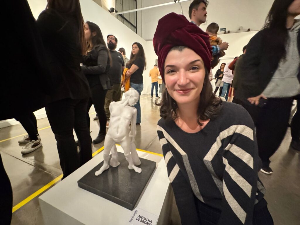A imagem mostra uma mulher sorrindo para a câmera, usando um turbante vinho na cabeça e um suéter escuro com listras brancas e cinzas. Ela está em uma galeria de arte, com outras pessoas ao fundo e uma escultura em primeiro plano. A escultura, em tons de branco, parece representar uma figura humana em uma pose contorcida. Há uma placa de identificação na base da escultura com o texto "MEDALHA DE BRONZE". O ambiente é iluminado e o chão é reflexivo, mostrando o reflexo das pessoas e da iluminação.