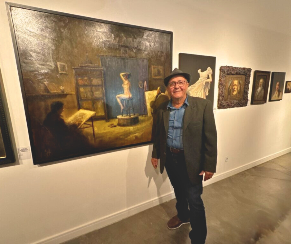  Homem de chapéu e óculos posa sorridente ao lado de um grande quadro que retrata um ateliê com modelo posando e artista desenhando.