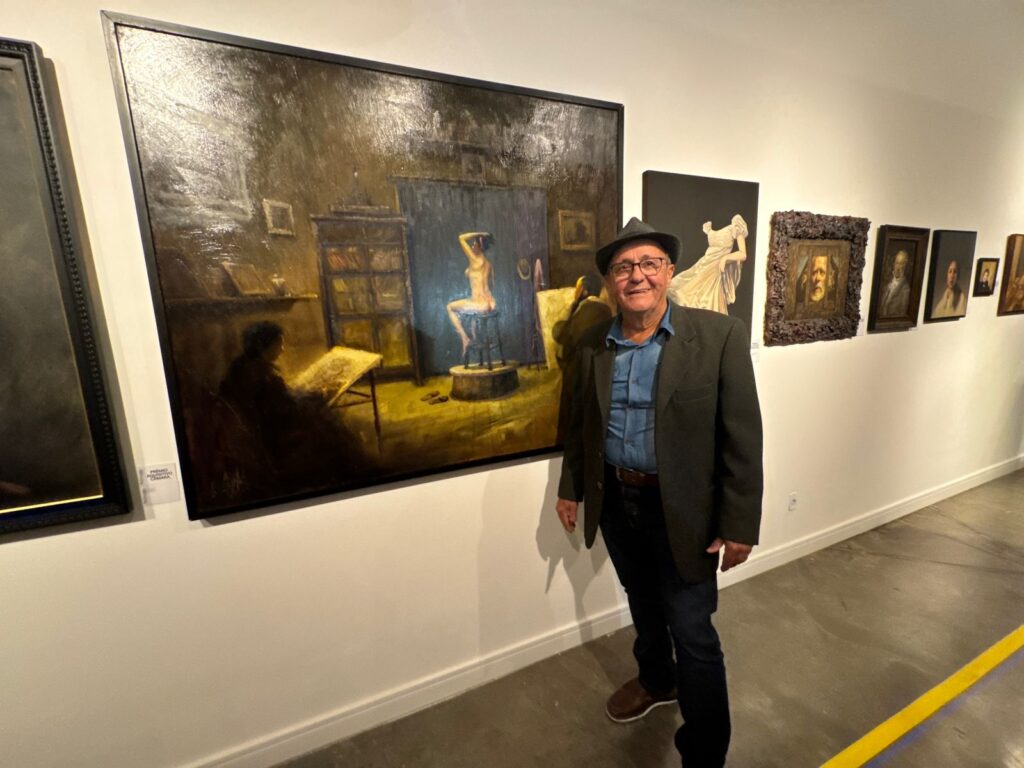 A imagem mostra um homem mais velho, sorrindo e posando em frente a uma grande pintura em uma galeria de arte. Ele está usando um chapéu, óculos, uma camisa jeans, um blazer escuro e calças. A pintura atrás dele é uma cena de um estúdio de arte, com uma figura feminina nua sentada em um pedestal, iluminada por um foco de luz. Há também uma figura sombreada sentada em uma cadeira, parecendo desenhar ou pintar. Ao lado dessa pintura principal, há outras obras de arte expostas na parede, incluindo retratos. O chão da galeria é de concreto e há uma linha amarela pintada no chão que se estende pela sala. A iluminação na galeria é suave, destacando as pinturas.