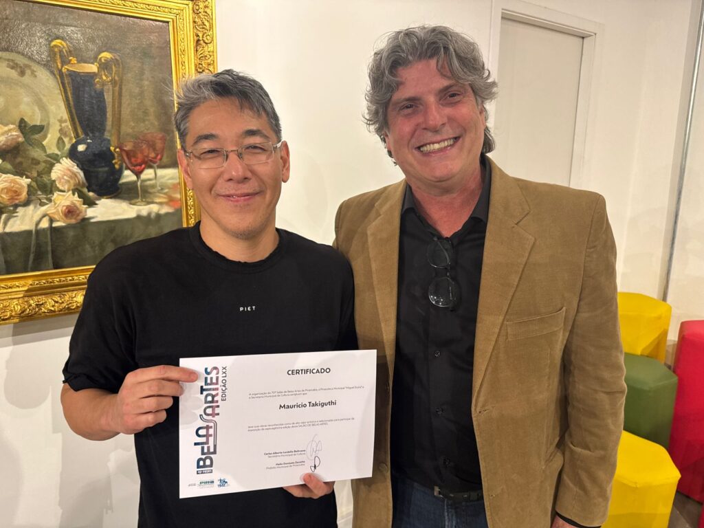 A imagem mostra dois homens sorrindo e posando para uma foto. O homem da esquerda, que parece ser de ascendência asiática, está vestindo uma camiseta preta com a palavra "PIET" em branco. Ele está segurando um certificado com a mão esquerda. O homem da direita, que tem cabelos grisalhos e usa um blazer marrom sobre uma camisa preta, está com o braço direito sobre o ombro do homem da esquerda. Atrás deles, há uma pintura emoldurada com um vaso e flores, e ao fundo, à direita, há alguns objetos coloridos em forma de cubo. O certificado que o homem da esquerda segura tem o título "CERTIFICADO" e menciona "BELA ARTES EDIÇÃO LXX", além do nome "Mauricio Takiguthi". Há também assinaturas e nomes de autoridades no certificado.