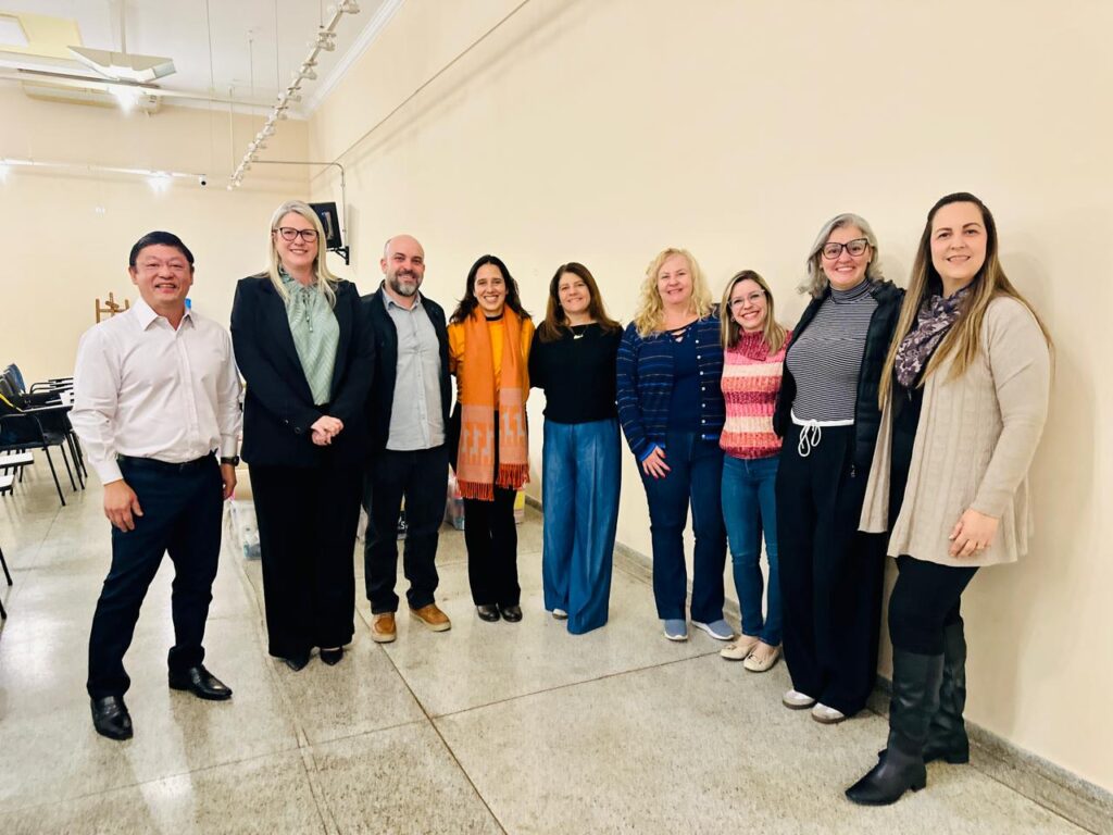 A imagem mostra um grupo de oito pessoas em pé em uma sala de aparência simples, com paredes claras e piso de azulejo. Elas estão vestidas de maneira casual e semi-formal, e ao fundo há mesas e cadeiras, sugerindo um ambiente de encontro ou reunião. A iluminação é forte, com luzes visíveis no teto.