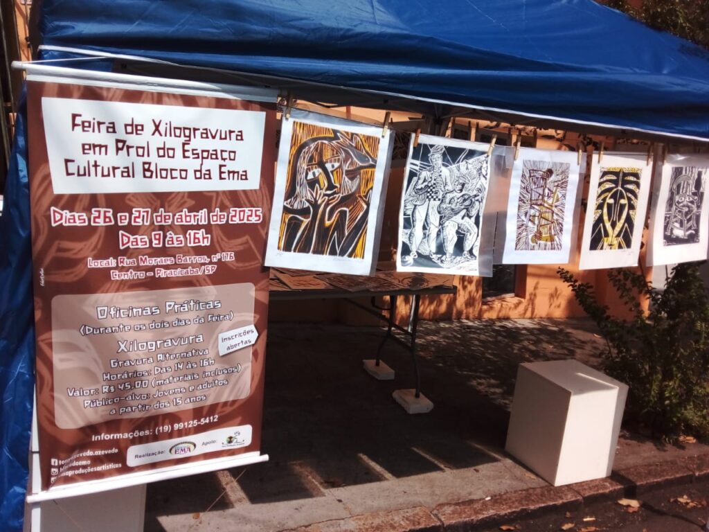 Varal de cordas com várias xilogravuras penduradas em folhas brancas, exibindo imagens de inspiração popular, em preto e amarelo. Ao lado, banner anuncia a Feira de Xilogravura em prol do Espaço Cultural Bloco da Ema.