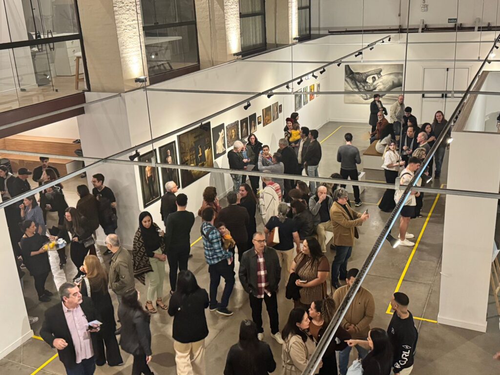 A imagem mostra um evento em uma galeria de arte, com muitas pessoas circulando e observando as obras expostas. A perspectiva é de cima, dando uma visão geral do espaço e da movimentação.As paredes da galeria estão repletas de quadros, com iluminação focada em cada peça. Há também uma grande obra de arte em destaque em uma das paredes, possivelmente o ponto focal da exposição.As pessoas presentes vestem-se de forma casual e variada, indicando um evento social e cultural. Algumas estão em grupos conversando, outras observam as obras individualmente. Há uma mesa com bebidas e aperitivos em um dos cantos, sugerindo um coquetel de abertura ou um evento de networking.O ambiente parece moderno e bem iluminado, com uma grande área aberta que permite a circulação livre dos visitantes. As linhas amarelas no chão podem indicar um caminho a ser seguido ou delimitar áreas específicas.