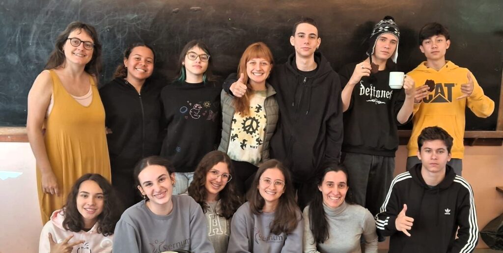 Grupo de treze pessoas posa sorrindo para a foto em frente a um quadro-negro. Na fileira de trás, estão em pé sete jovens e duas mulheres adultas. Na frente, sentados ou agachados, há quatro adolescentes. Alguns fazem sinal de positivo ou hang loose com as mãos. Dois jovens seguram caneca e cookie. Todos estão vestidos de forma casual, com moletons, camisetas e roupas confortáveis. O clima é descontraído, de confraternização em ambiente escolar.