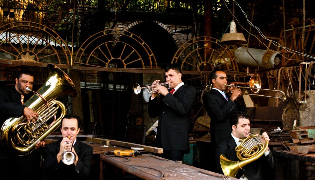 Cinco músicos do grupo BrassUka, vestidos com ternos pretos, gravatas e camisas claras, tocam seus instrumentos de sopro em meio a um ambiente industrial, com estruturas enferrujadas, rodas metálicas e ferramentas. A cena remete a um contraste entre a música erudita e o cenário fabril, reforçando o estilo irreverente do grupo.