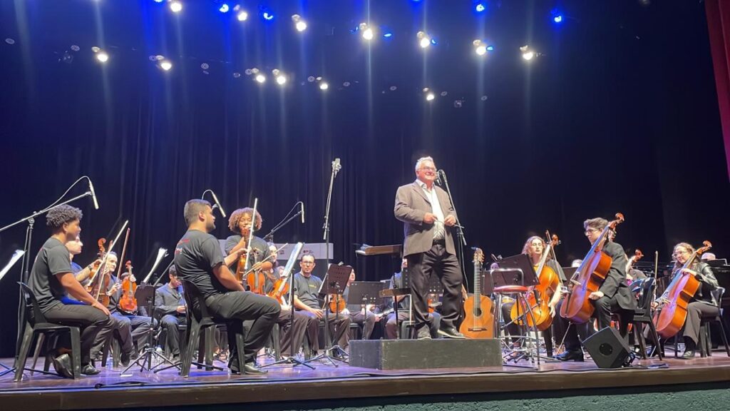 Maestro sorri em pé no centro do palco, cercado por músicos de orquestra com instrumentos de corda, durante apresentação iluminada por refletores.
