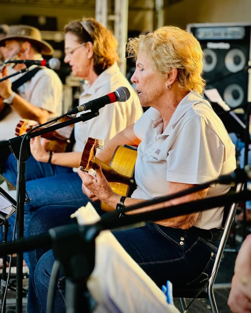 Uma senhora loira, de cabelos curtos, está sentada e canta enquanto toca viola caipira em frente a um microfone. Ao seu lado, outras mulheres, também com camisas claras, tocam e cantam. A cena transmite emoção e concentração durante a apresentação.