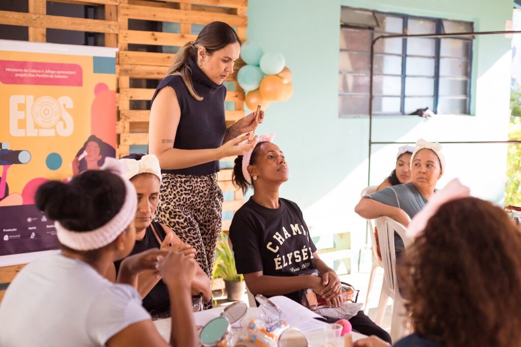 Uma mulher, vestindo um top sem mangas e calças estampadas de onça, está demonstrando uma técnica de beleza para um grupo de mulheres. Ela segura um pequeno aplicador perto do rosto de uma mulher sentada à sua frente, que usa uma camiseta preta com "CHAMPION ÉLYSÉE Paris 1970" estampada e uma faixa rosa na cabeça.Várias outras mulheres também estão sentadas, algumas usando faixas, e parecem estar participando ou observando a demonstração. O ambiente parece ser um workshop ou aula informal, com um banner ao fundo que diz "Ministério da Cultura e Jofege apresentam projeto Elos Partilha de Saberes" e a palavra "ELOS" em destaque. A atmosfera geral sugere um evento comunitário focado em aprender e compartilhar habilidades.