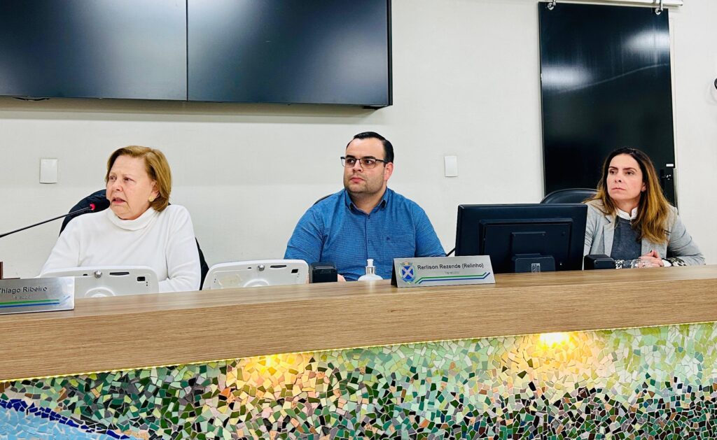 A imagem mostra três pessoas sentadas atrás de uma mesa de madeira com um mosaico colorido na frente. A pessoa mais à esquerda é uma mulher mais velha com cabelo loiro curto, vestindo um suéter branco de gola alta. Ela está falando e olhando para a esquerda, com um microfone à sua frente. A pessoa no meio é um homem com cabelo escuro curto, usando óculos e uma camisa azul de botões. Ele está olhando para a direita. À sua frente, há uma placa com o nome "Rerlison Rezende (Relinho)" e "Vereador", juntamente com um brasão. A pessoa mais à direita é uma mulher com cabelo castanho comprido, vestindo um blazer cinza sobre uma blusa escura. Ela está olhando para a frente, com as mãos apoiadas na mesa. Há um monitor de computador à sua frente. O ambiente parece ser uma sala de reuniões ou um auditório, com uma tela escura ao fundo e uma parede branca.