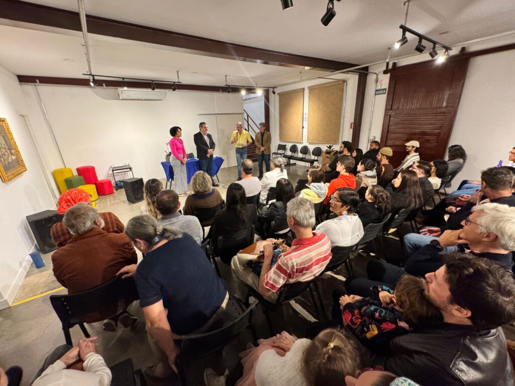 A imagem retrata um evento em um espaço interno, possivelmente uma galeria de arte ou um centro cultural, com um público sentado em cadeiras voltado para um palco improvisado. No palco, três pessoas estão de pé, com duas mulheres e um homem em destaque. Uma mulher, vestindo uma blusa rosa vibrante e calças cinzas, está ao lado de um homem de terno escuro. Um terceiro homem, vestindo uma camisa amarela, está um pouco mais afastado, segurando um papel.O ambiente é iluminado por luzes de teto e possui uma parede branca ao fundo, onde estão pendurados alguns objetos decorativos, como quadros e almofadas coloridas (vermelha, amarela e verde). Há também equipamentos de som e caixas acústicas no chão, sugerindo uma apresentação ou palestra. O público, composto por cerca de trinta pessoas, está variadamente vestido, com a maioria olhando na direção do palco. A atmosfera parece ser de atenção e engajamento com o que está acontecendo à frente.