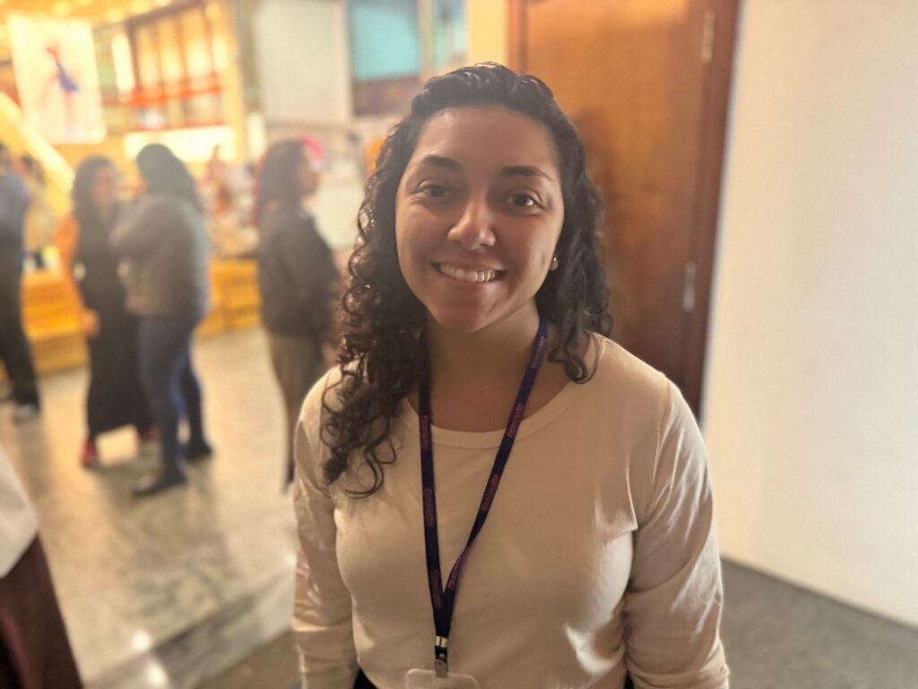 A imagem mostra uma mulher sorrindo para a câmera. Ela tem cabelo escuro e cacheado e usa uma blusa de manga comprida de cor clara. Um cordão azul com um crachá visível está pendurado em seu pescoço. O fundo está desfocado, mas parece ser um ambiente interno com outras pessoas e uma porta de madeira.