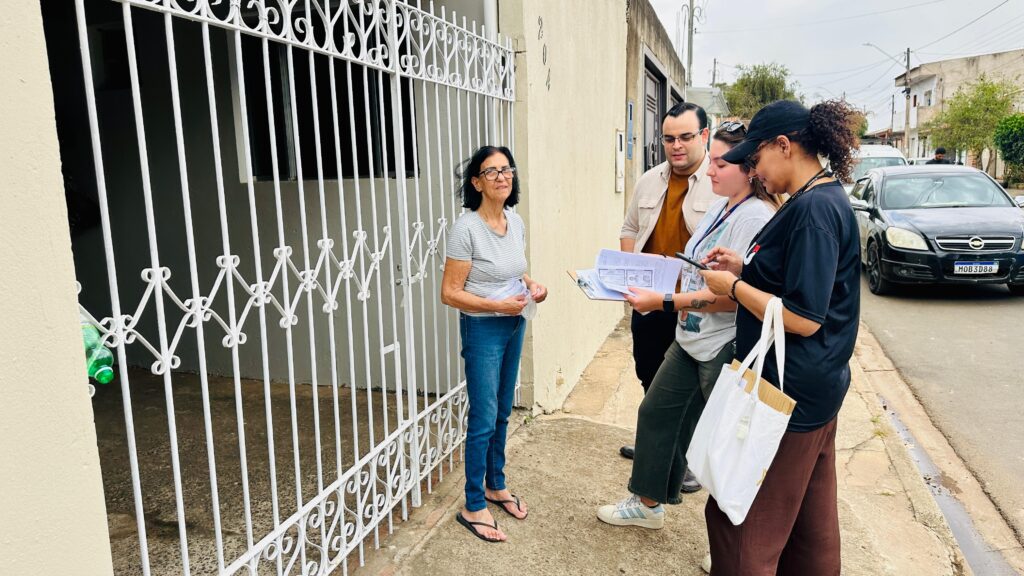 A imagem mostra uma cena externa em frente a um portão branco de metal decorado, com o número “204” visível na parede. Quatro pessoas estão envolvidas: uma está dentro do portão, vestindo camiseta cinza e jeans, enquanto outras três estão do lado de fora, segurando papéis e um celular, aparentemente em diálogo com quem está dentro. Há um carro preto estacionado próximo, com a placa visível, e o ambiente parece ser de um bairro residencial. A interação sugere uma visita técnica, entrega de documentos ou coleta de informações, com clima de cordialidade e atenção.