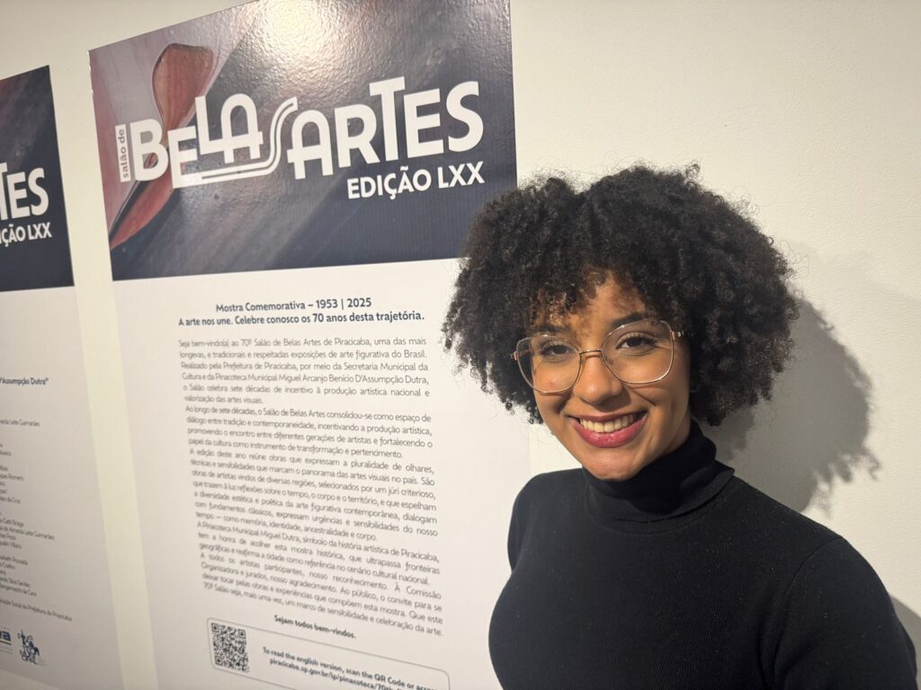 A imagem mostra uma mulher sorrindo para a câmera, com um pôster de arte ao fundo. O pôster anuncia a 70ª Edição do Salão de Belas Artes de Piracicaba, que celebra 70 anos de história. O texto no pôster descreve o evento como uma das exposições de arte figurativa mais antigas e tradicionais do Brasil, realizado pela Secretaria Municipal da Cultura e pela Pinacoteca Municipal Miguel Arcanjo Benício D'Assumpção Dutra. O evento reúne obras que expressam a pluralidade de olhares, técnicas e sensibilidades das artes visuais do país, com obras de artistas de diversas regiões selecionadas por um júri criterioso. O pôster também convida o público a celebrar a arte e a sensibilidade.