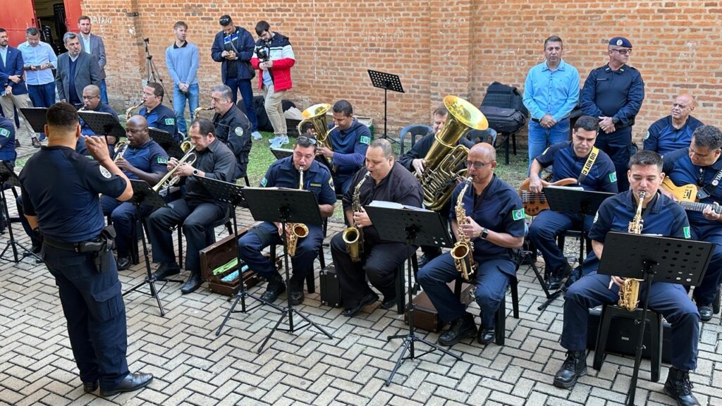 Integrantes de uma banda musical vestindo uniformes azul escuro se apresentam com seus instrumentos musicais, em um espaço livre. A frente aparece um senhor com uma partitura, regendo os músicos. Atrás da banda aparecem algumas pessoas, umas olhando no celular e outras prestando atenção na apresentação.