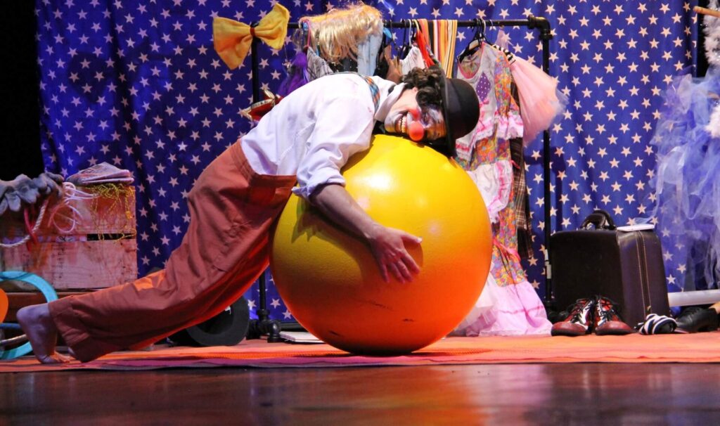 O palhaço interage de forma divertida com uma grande bola amarela, que ele abraça deitado sobre o palco. Ao fundo, um tecido azul com estrelas brancas cobre parte do cenário, e há cabides com fantasias e adereços coloridos.