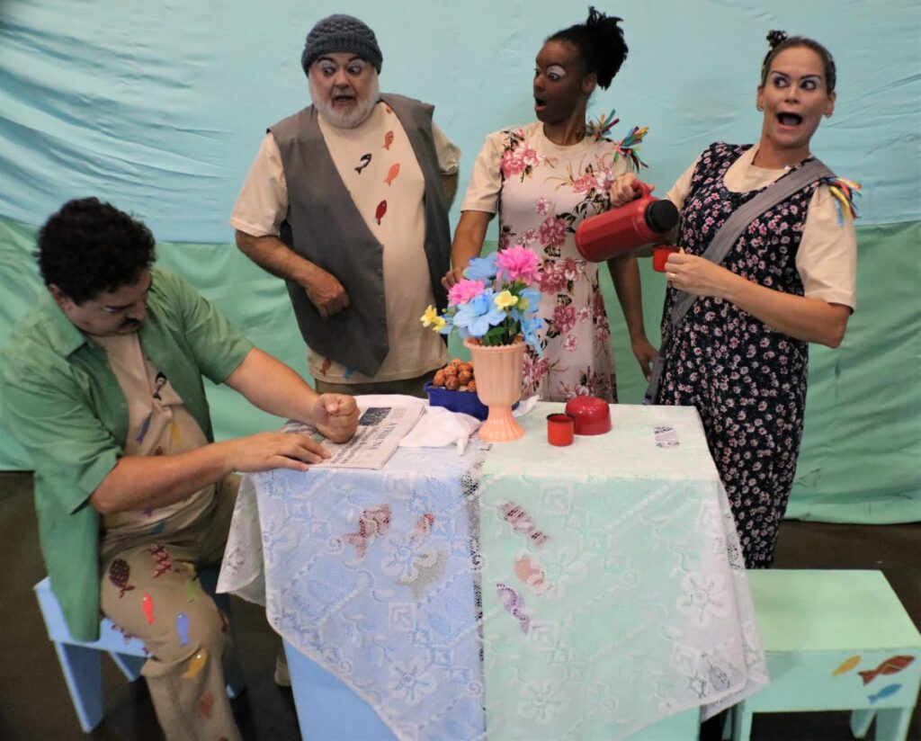 Quatro atores estão reunidos ao redor de uma mesa coberta com toalha rendada branca. Um deles, sentado, bate com força no jornal sobre a mesa. Outro, de pé, observa surpreso. Uma atriz com vestido floral serve café com expressão exagerada de espanto, enquanto a outra também reage com surpresa. Ao centro da mesa há um vaso com flores coloridas e um prato de quitutes. As roupas e expressões remetem ao estilo cômico e caipira.