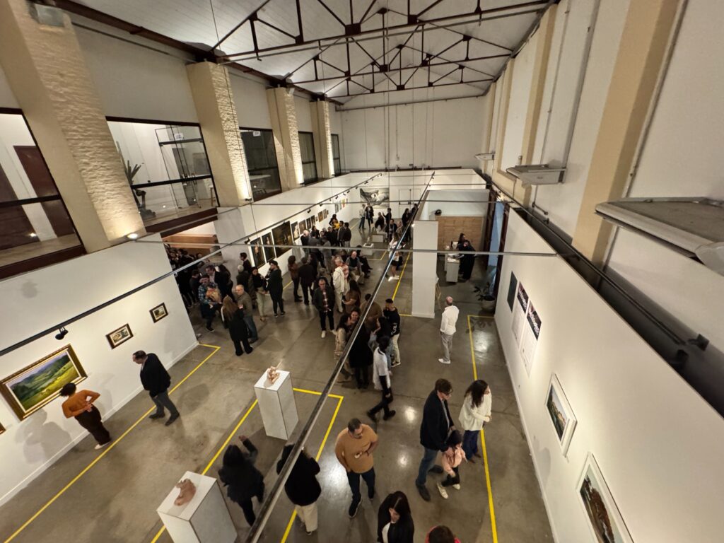 Salão de arte com público caminhando e observando quadros e esculturas em exposição.