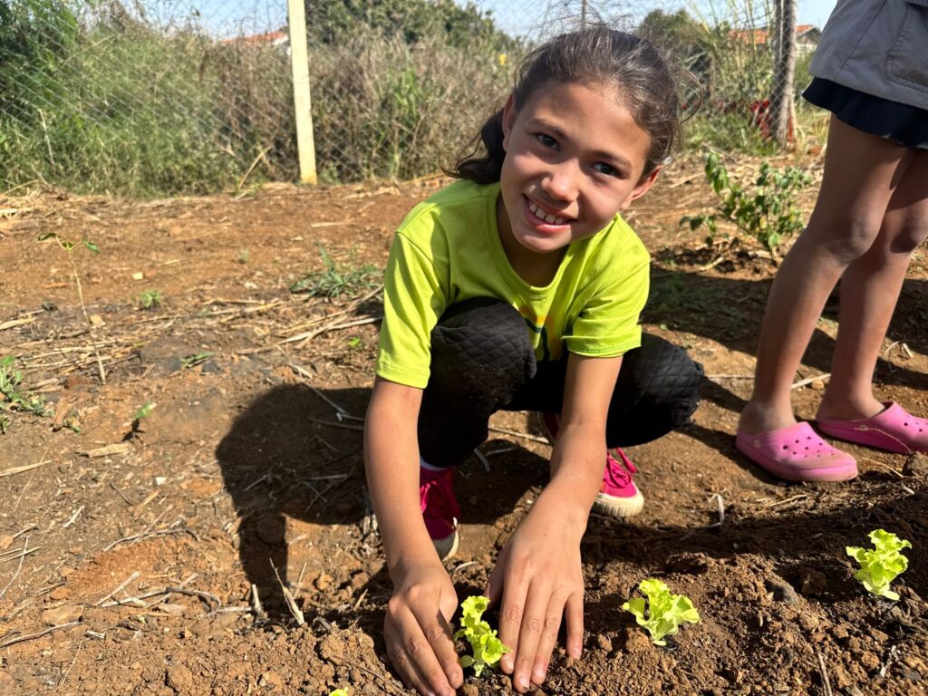 A imagem mostra duas crianças em uma área de plantio ao ar livre. Uma está ajoelhada e com foco total em plantar pequenas mudas de alface na terra seca, usando as mãos com cuidado. A outra criança está em pé ao lado, observando a ação, com sapatos rosa chamativos que contrastam com o solo e a vegetação ao redor. O local é cercado por uma cerca simples e apresenta alguns sinais de uso frequente, como plantas dispersas e partes do solo remexidas.