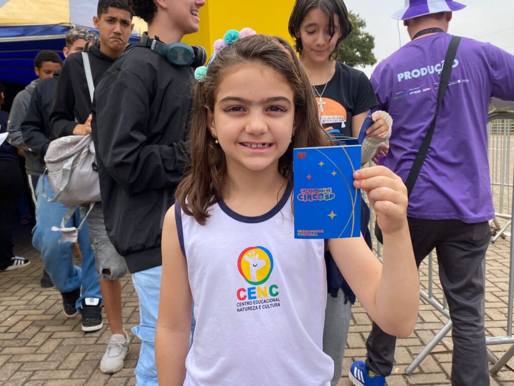 Menina de aproximadamente 8 anos sorri para a câmera segurando o “Passaporte Cultural” azul do Festival de Circo SP. Ela veste camiseta branca com logo do Centro Educacional Natureza e Cultura (CENC).