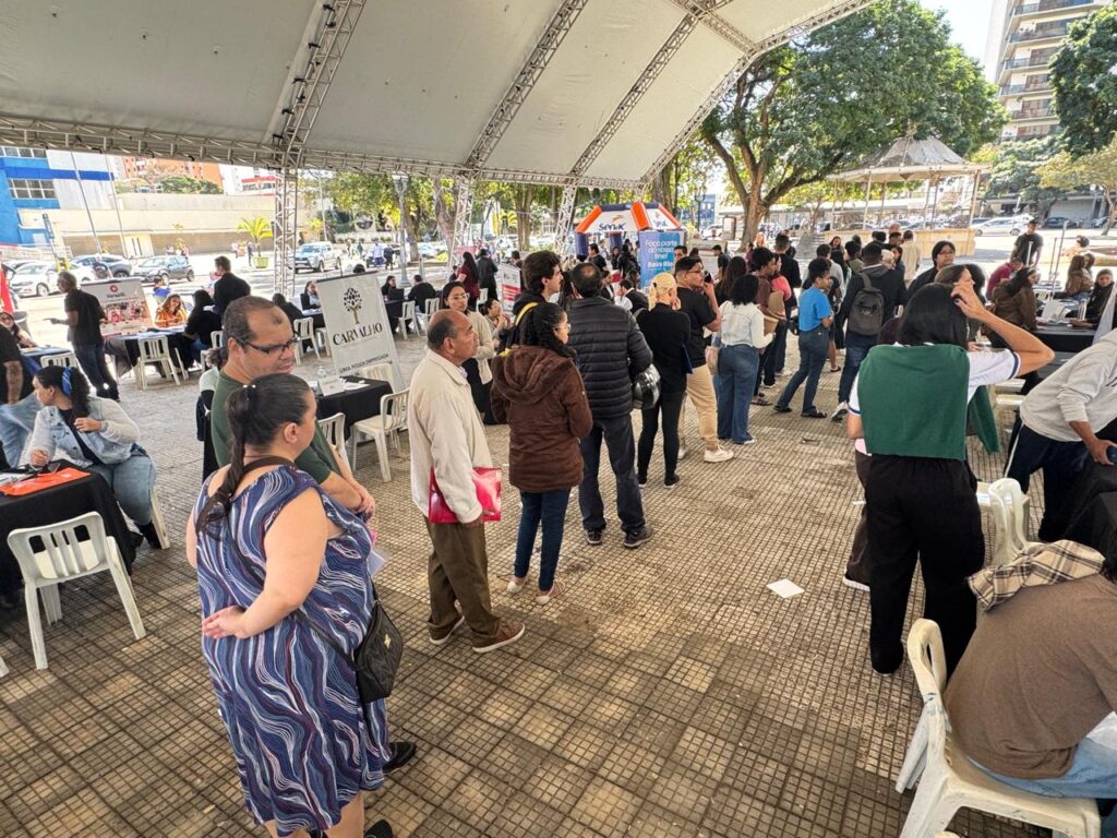 A imagem mostra um evento ao ar livre, possivelmente uma feira de empregos ou um evento comunitário, sob uma grande tenda branca. Há uma multidão de pessoas em pé e sentadas, interagindo e se movendo pelo espaço. Várias mesas com materiais informativos e banners estão dispostas, com algumas pessoas em pé atrás delas, possivelmente representando organizações ou oferecendo informações. O ambiente parece ser urbano, com edifícios e árvores visíveis ao fundo. A iluminação sugere que é dia, com luz solar entrando pela lateral da tenda.