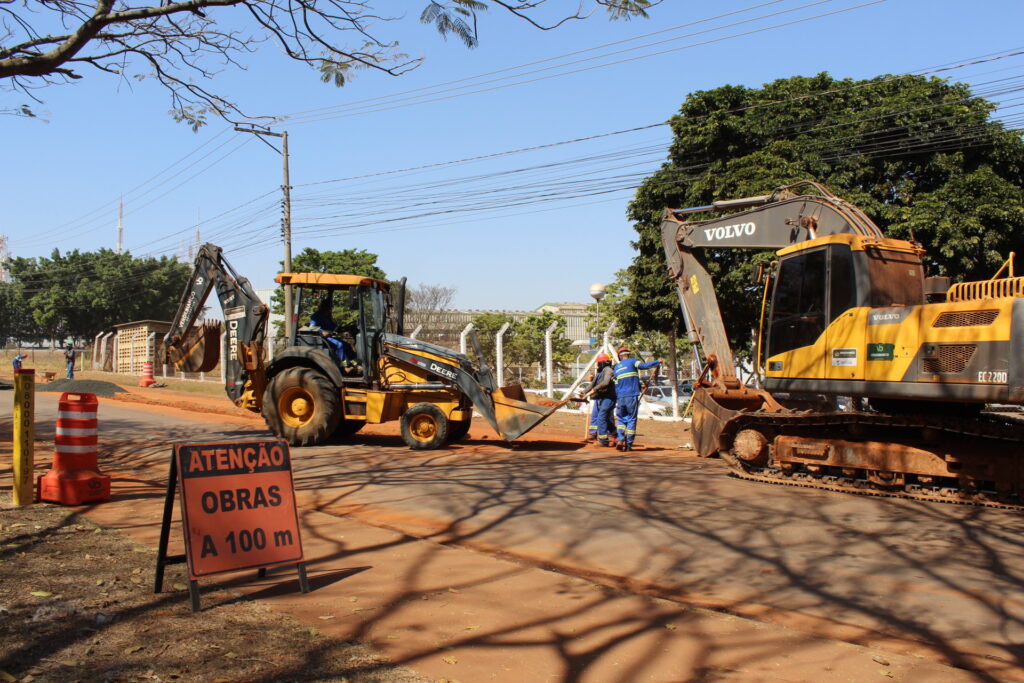 A imagem mostra uma cena de obras em andamento, com dois grandes equipamentos de construção e trabalhadores. À esquerda, um retroescavadeira amarela da marca John Deere está em operação. Um trabalhador está visível na cabine, operando a máquina. A caçamba da retroescavadeira está levantada e parece estar em processo de escavação ou movimentação de terra. À direita, uma escavadeira maior, da marca Volvo, também amarela, está estacionada. A marca "VOLVO" é claramente visível na parte superior da cabine. Abaixo da marca Volvo, há outros logotipos e textos, incluindo "EC 2200". A escavadeira Volvo também parece estar em um local de trabalho, com sua lança e caçamba posicionadas. No centro da imagem, entre as duas máquinas, três trabalhadores vestindo uniformes azuis e capacetes (um laranja, um azul e um cinza) estão reunidos. Eles parecem estar discutindo ou supervisionando o trabalho. Um deles está segurando uma ferramenta, possivelmente uma pá. Um sinal de trânsito em forma de cavalete está posicionado em primeiro plano, à esquerda. O sinal tem um fundo laranja e texto preto que diz "ATENÇÃO OBRAS A 100 m", indicando a proximidade de uma área de obras. Ao lado do sinal, há cones de trânsito laranja e branco, e um poste com uma placa numerada. O chão ao redor é de terra avermelhada, sugerindo que o local está em processo de movimentação de terra ou pavimentação. A cena é iluminada por uma luz solar forte, criando sombras distintas no chão, inclusive as sombras de galhos de árvores. Ao fundo, há uma linha de postes de eletricidade com vários fios, árvores verdes e algumas construções. O céu está limpo e azul. A atmosfera geral é de um dia de trabalho ativo em um canteiro de obras.