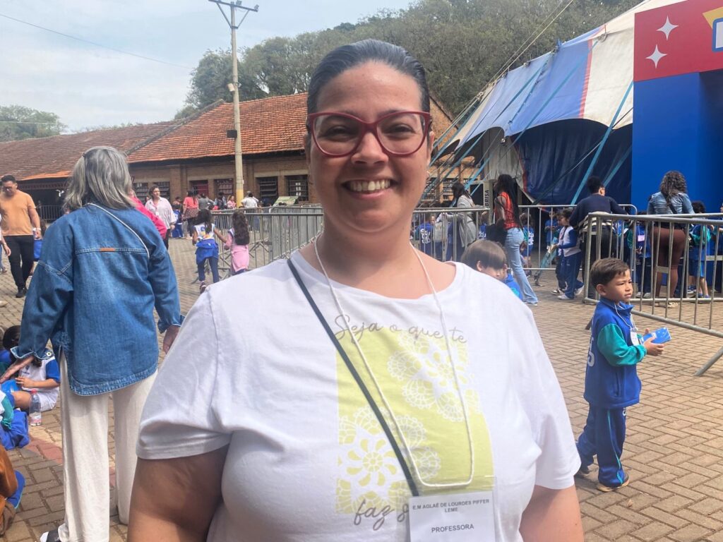 Mulher de óculos vermelhos e cabelo preso sorri em frente à entrada de uma lona de circo. Ela veste camiseta branca com estampa amarela e um crachá que a identifica como professora da Escola Municipal Prof.ª Aglaé de Lourdes Piffer Leme.