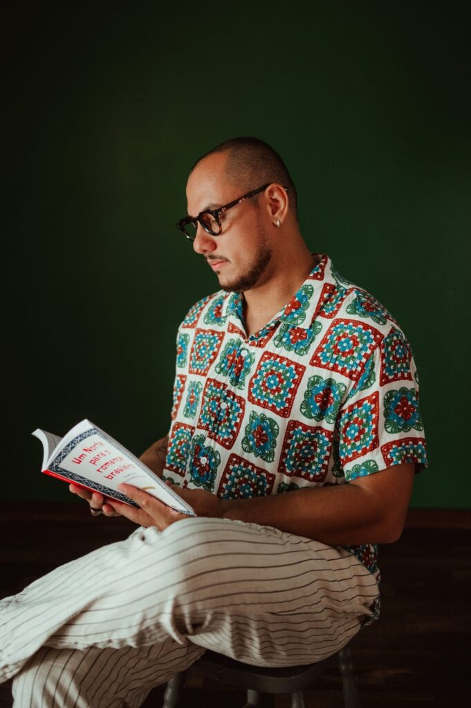 Homem careca, de óculos e barba rala, lê um livro enquanto está sentado de lado em um banco. Ele veste uma camisa estampada colorida em tons de verde, vermelho e azul, e calça clara listrada. Ao fundo, a parede é verde-escura, criando contraste com a cena iluminada.