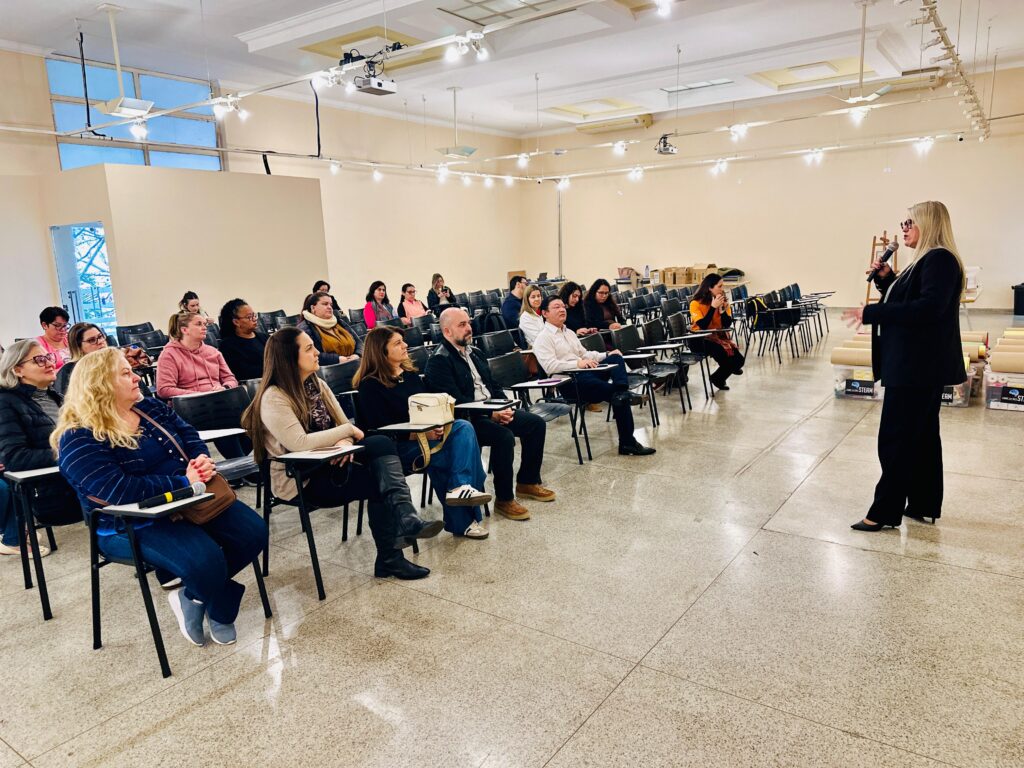 A imagem mostra uma sala de aula ou ambiente de seminário, onde um grupo de pessoas está sentado em cadeiras com pranchetas, voltado para um palestrante que fala ao microfone. O espaço é bem iluminado e há materiais empilhados ao fundo, como caixas e rolos de papel. O público parece atento à apresentação.