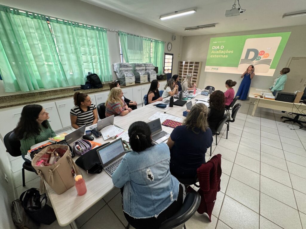 A imagem mostra um ambiente de formação ou reunião pedagógica, com foco em avaliações externas. Um grupo de pessoas está reunido em torno de uma mesa, utilizando laptops, cadernos e outros materiais de apoio. Duas pessoas estão em pé à frente da sala, conduzindo a apresentação exibida em uma tela com o título “DIA D Avaliações externas”. O espaço é organizado, com cortinas verdes, armários brancos e um relógio na parede, sugerindo um ambiente escolar.
