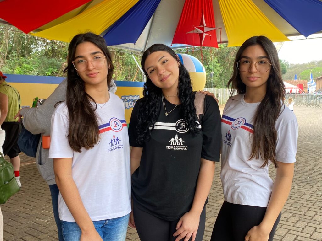 Três meninas adolescentes, de cabelos longos e soltos, posam sorridentes em frente ao espaço do festival. Duas delas usam camisetas brancas da Escola Estadual com a palavra “Integral” estampada, e a do meio veste camiseta preta.