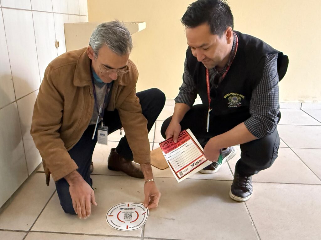 dois homens estão agachados colando adesivo para tampar ralos e evitar criadouros da dengue