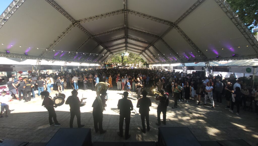 A imagem mostra um grande evento ao ar livre, possivelmente um festival ou feira, sob uma tenda branca imponente. A tenda é sustentada por uma estrutura metálica complexa e iluminada por luzes roxas que criam um ambiente vibrante.Uma multidão considerável de pessoas está reunida sob a tenda, com a maioria delas em pé e interagindo. Há uma banda tocando na frente, com músicos vestidos de forma semelhante, alguns tocando instrumentos de sopro e percussão. A banda está posicionada em um palco ou área elevada, com caixas de som visíveis.O ambiente parece ser diurno, com luz solar entrando nas laterais da tenda, criando sombras interessantes no chão. Ao fundo, é possível ver árvores e um céu azul, sugerindo que o evento está ocorrendo em um espaço aberto, como uma praça ou parque. Há também algumas barracas ou estandes com logotipos e informações visíveis nas laterais da área coberta. A atmosfera geral é de celebração e entretenimento.