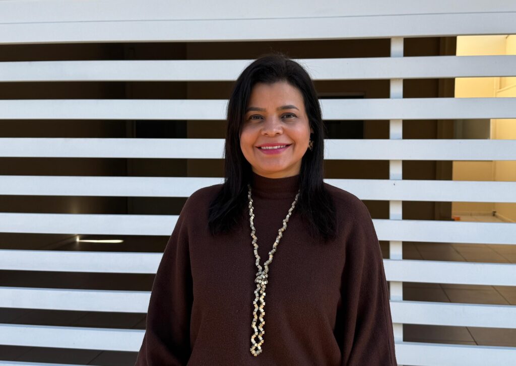 a imagem mostra uma mulher de pele parda, cabelo pretos lisos, sorridente e usando blusa marrom e colar prateado posa para fotografia em frente a uma residência com portão branco