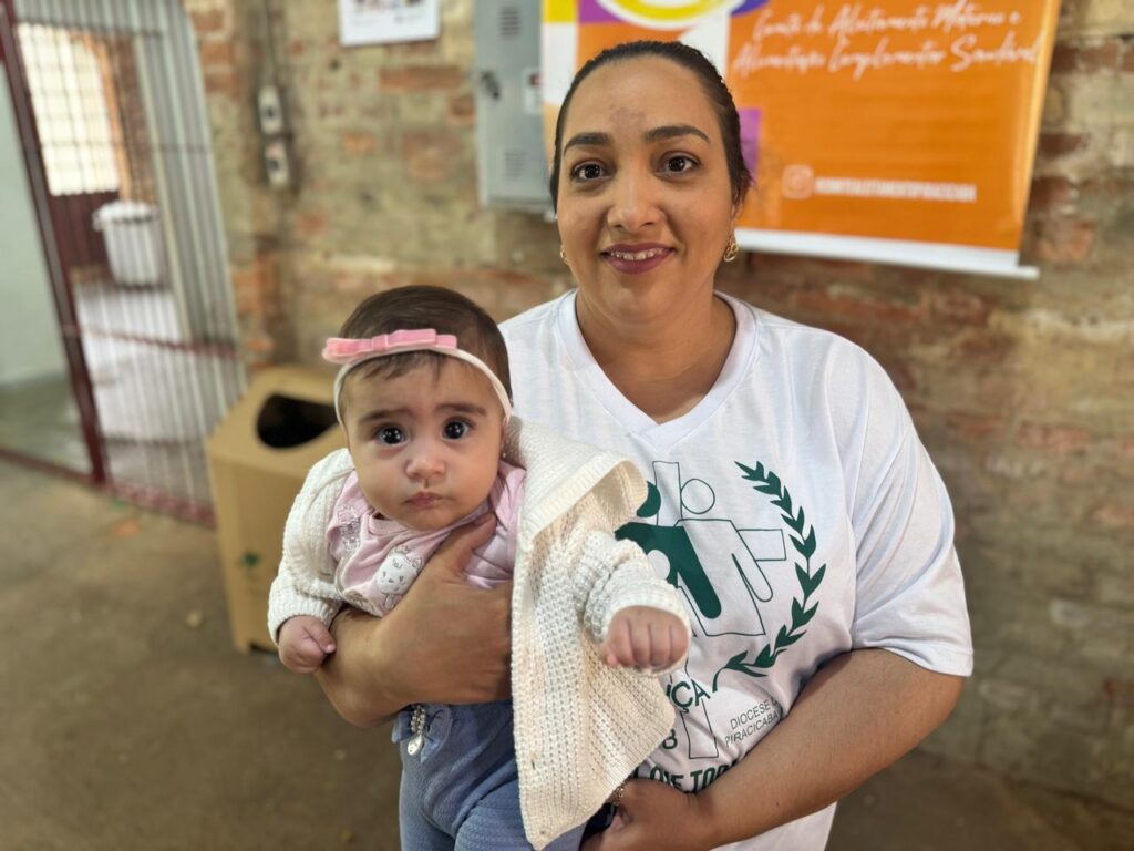 Uma mulher segura um bebê no colo. A mulher está usando uma camiseta branca com um logotipo verde. O bebê está usando uma faixa rosa na cabeça e um suéter branco. Ambos estão sorrindo para a câmera. O fundo é uma parede de tijolos e um banner laranja com texto em português.