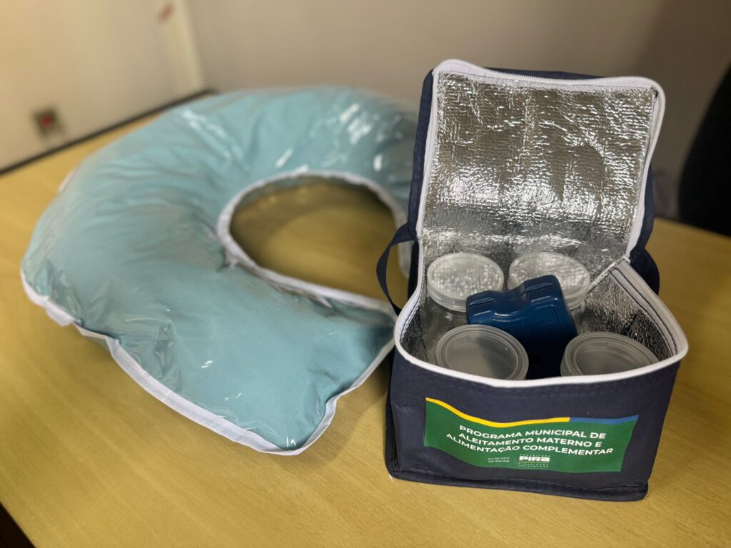A imagem mostra um kit para amamentação, composto por uma almofada de amamentação e uma bolsa térmica. A almofada de amamentação é grande e em formato de "C", com uma cor azul clara e uma capa plástica transparente. Ela parece ser macia e confortável, ideal para apoiar o bebê durante a amamentação. Ao lado da almofada, há uma bolsa térmica azul escura, com o interior forrado com material isolante prateado. A bolsa está aberta, revelando quatro potes de armazenamento transparentes com tampas brancas e um objeto azul, que ajuda a manter a temperatura da bolsa térmica. Na parte frontal da bolsa, há uma etiqueta com as inscrições "PROGRAMA MUNICIPAL DE ALEITAMENTO MATERNO E ALIMENTAÇÃO COMPLEMENTAR", "SECRETARIA DE SAÚDE" e "PIRACICABA", indicando que o kit faz parte de uma iniciativa de saúde pública da cidade de Piracicaba.