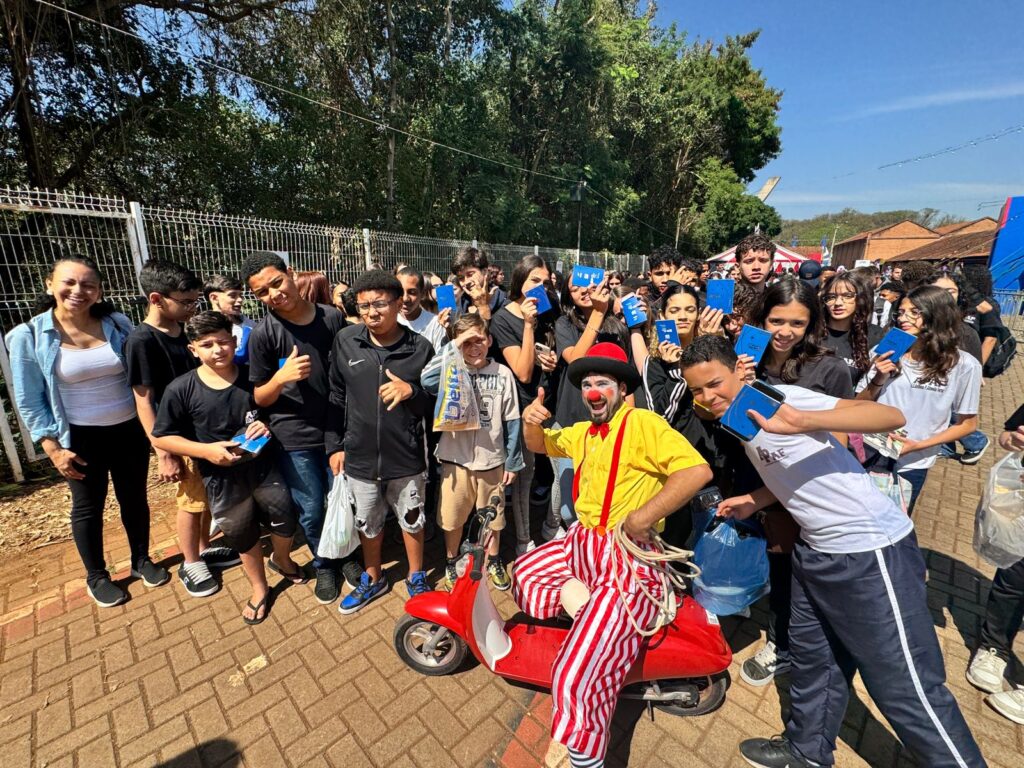 Pra cego ver: Grupo de adolescentes posa ao ar livre ao lado de um palhaço que segura um mini triciclo vermelho. Os jovens mostram na mão o passaporte azul do Festival de Circo. Todos sorriem para a foto.