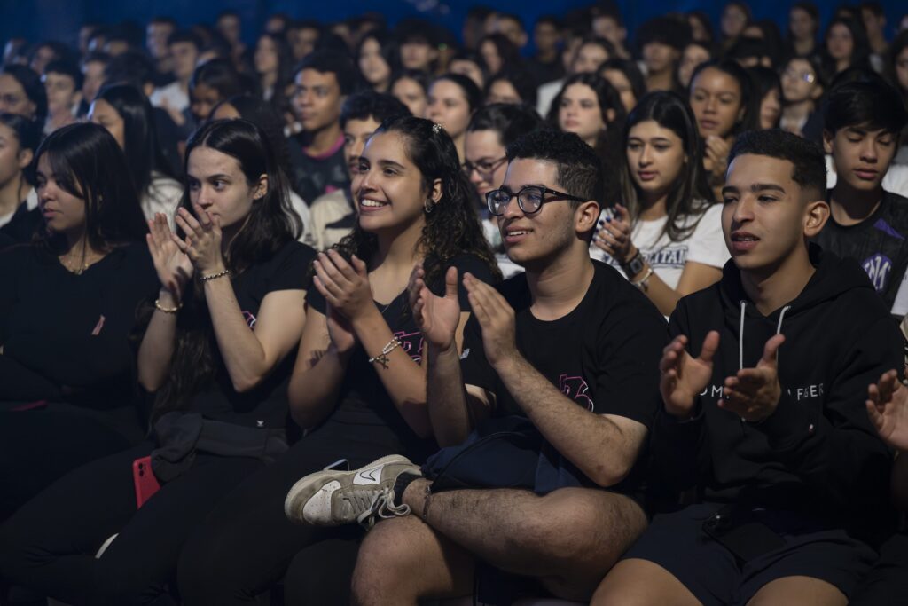Jovens vestidos de preto e branco, sentados em cadeiras, aplaudem com entusiasmo durante apresentação dentro da lona do festival.
