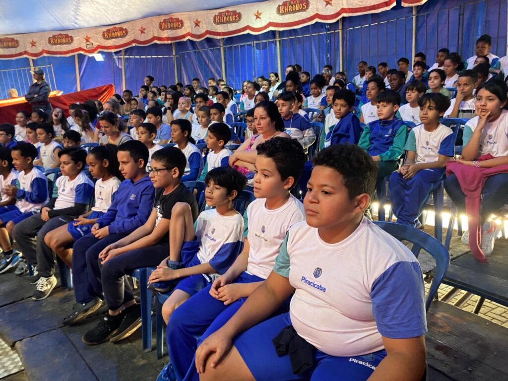 Vista ampla de dentro da lona do circo, mostrando centenas de crianças uniformizadas ocupando todas as arquibancadas. A cena transmite energia, diversidade e a grandiosidade do evento, com o espaço totalmente tomado pelo público