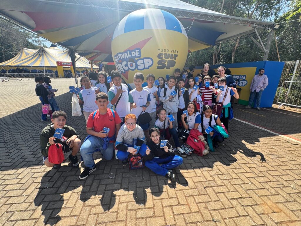 Grupo de estudantes do 5º ano posa em frente ao balão cenográfico do Festival de Circo SP. Eles seguram os passaportes azuis do festival e sorriem. Ao fundo, aparecem as lonas coloridas montadas no Engenho Central.