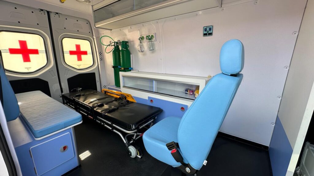 A imagem mostra o interior de uma ambulância, com foco nos equipamentos médicos e assentos. O ambiente é bem iluminado, com paredes brancas e detalhes em azul. Duas garrafas de oxigênio verdes estão montadas na parede, acompanhadas de medidores de pressão e recipientes para líquidos. Um leito de paciente, com cintos de segurança, está preparado para uso. Há também um assento azul claro em primeiro plano, com um cinto de segurança visível, e um assento mais baixo ao lado, acolchoado. As janelas grandes apresentam o símbolo da Cruz Vermelha, enquanto uma prateleira de vidro com itens coloridos e armários azuis com alças vermelhas completam o espaço. Um pequeno painel de controle com botões e luzes está fixado na parede superior direita.
