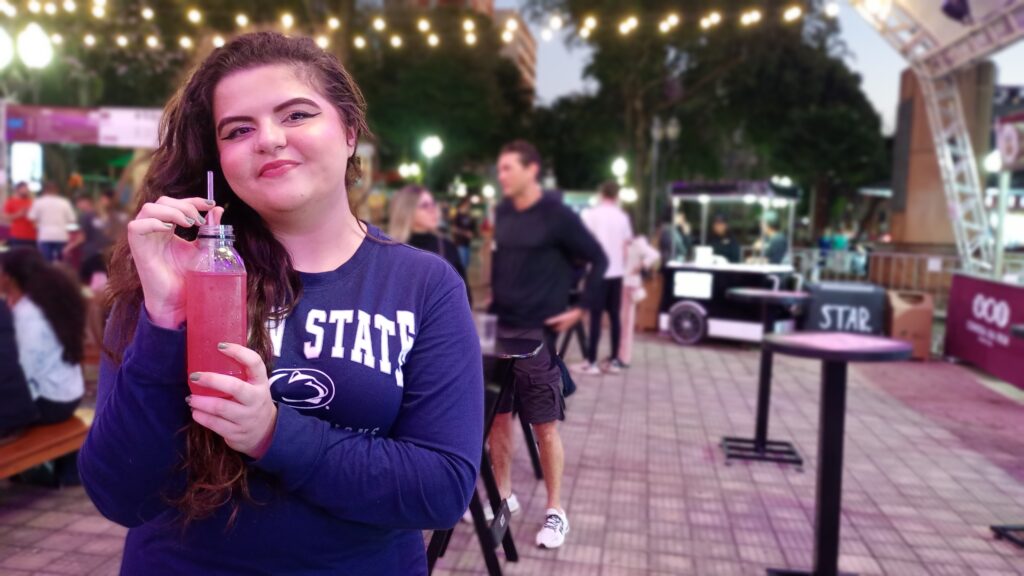 Uma jovem sorri para a câmera enquanto segura uma garrafa de plástico com uma bebida rosa e um canudo. Ela está usando uma blusa azul de manga comprida com "PENN STATE" escrito em branco no peito. O fundo está desfocado, mas mostra uma área externa com pessoas, luzes de corda penduradas e barracas. Há também mesas e cadeiras em primeiro plano, sugerindo um ambiente de evento ou festival.