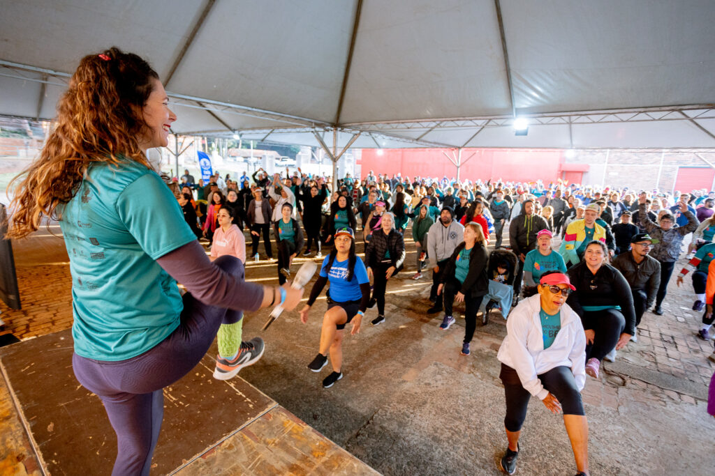 A imagem retrata um grupo de pessoas participando de uma aula de ginástica ao ar livre, sob uma grande tenda. No primeiro plano, uma instrutora sorridente, com cabelos castanhos ondulados e uma camiseta verde-água, está de pé em um palco de madeira, demonstrando um movimento com a perna levantada.O grupo de participantes, composto por homens e mulheres de diversas idades, está posicionado em um pátio cimentado, todos em pé e seguindo os movimentos da instrutora. A maioria veste roupas esportivas em tons variados, como azul, rosa, verde e preto. O fundo mostra uma estrutura de tenda branca e, mais atrás, uma parede vermelha e algumas árvores, sugerindo um ambiente externo. A iluminação indica que o evento está ocorrendo durante o dia.